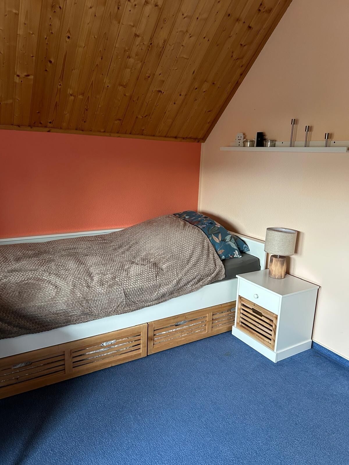Une chambre avec un plafond en bois, un lit recouvert d’une couverture à motifs, une table de chevet avec une lampe et une étagère contenant des ustensiles de cuisine.