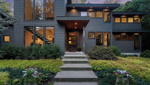 Une maison moderne à deux étages, avec un chemin en béton menant à l’entrée, entourée de parterres bien entretenus et de plantes en pot.