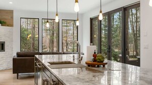 Une cuisine moderne dotée d’un grand îlot central, de luminaires suspendus et de grandes fenêtres offrant une vue sur la forêt.
