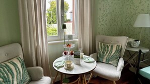A cozy room with a round table set for tea, two armchairs, a sofa, and a view through a window with curtains.