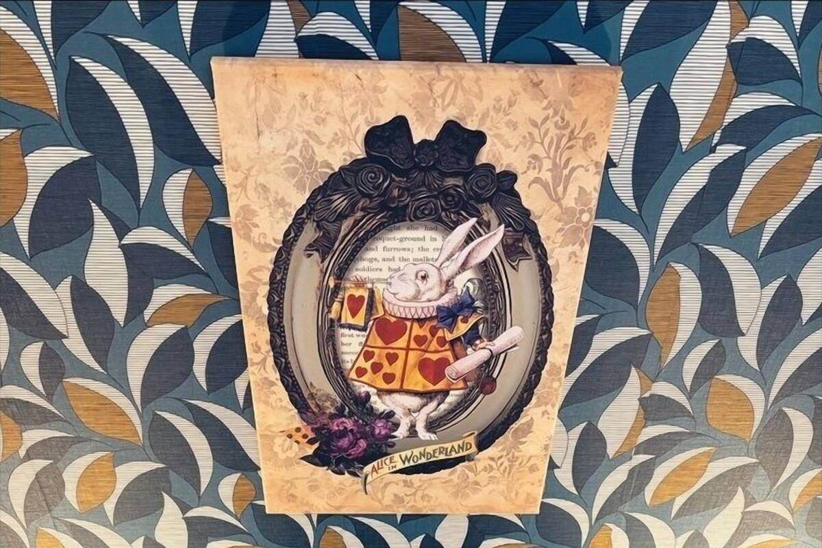 A wall with a patterned wallpaper and a framed picture of a dog wearing a dress with hearts. Below, there are pillows with designs of a cat and a character from a story.