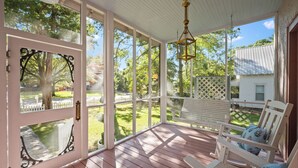 En overdækket veranda med trægulv, hvide gelændere og et hvidt hegn. Der er en gyngestol med blomsterpuder og en hængende lanterne.