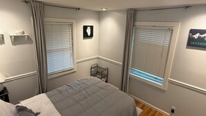 A bedroom with a bed, two windows with blinds, a wall-mounted shelf with a decorative item, and a framed picture.