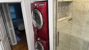 A laundry area with a red washer and dryer, a shower with a glass door, and a bedroom visible through an open door.