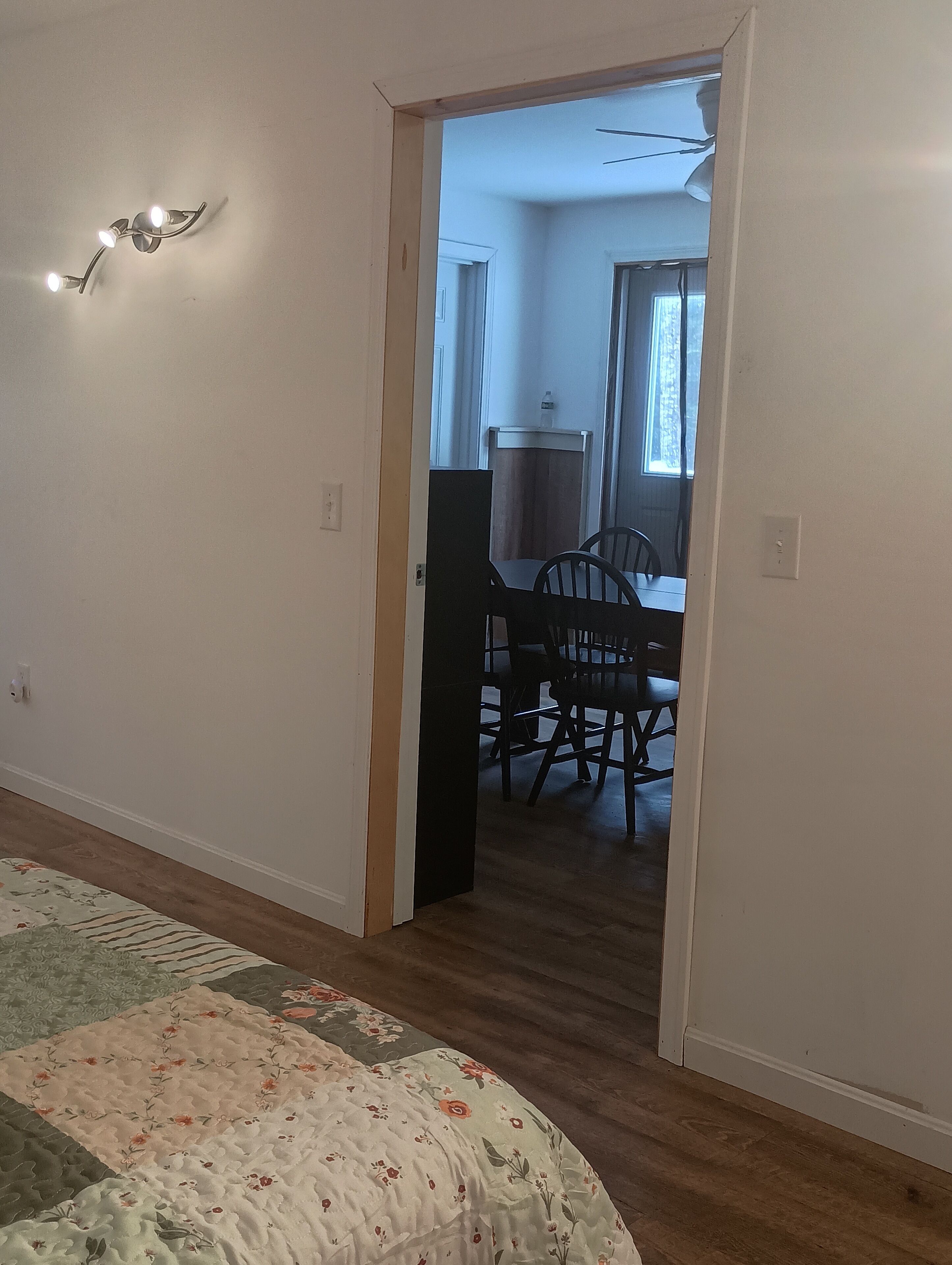 A room with a bed, a door leading to a dining area with a table and chairs, and a wall-mounted light fixture.