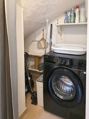 Ein kleiner Hauswirtschaftsraum mit einer Waschmaschine mit Frontlader, einem Regal mit Reinigungsmitteln und einem an die Wand gelehnten Besen.