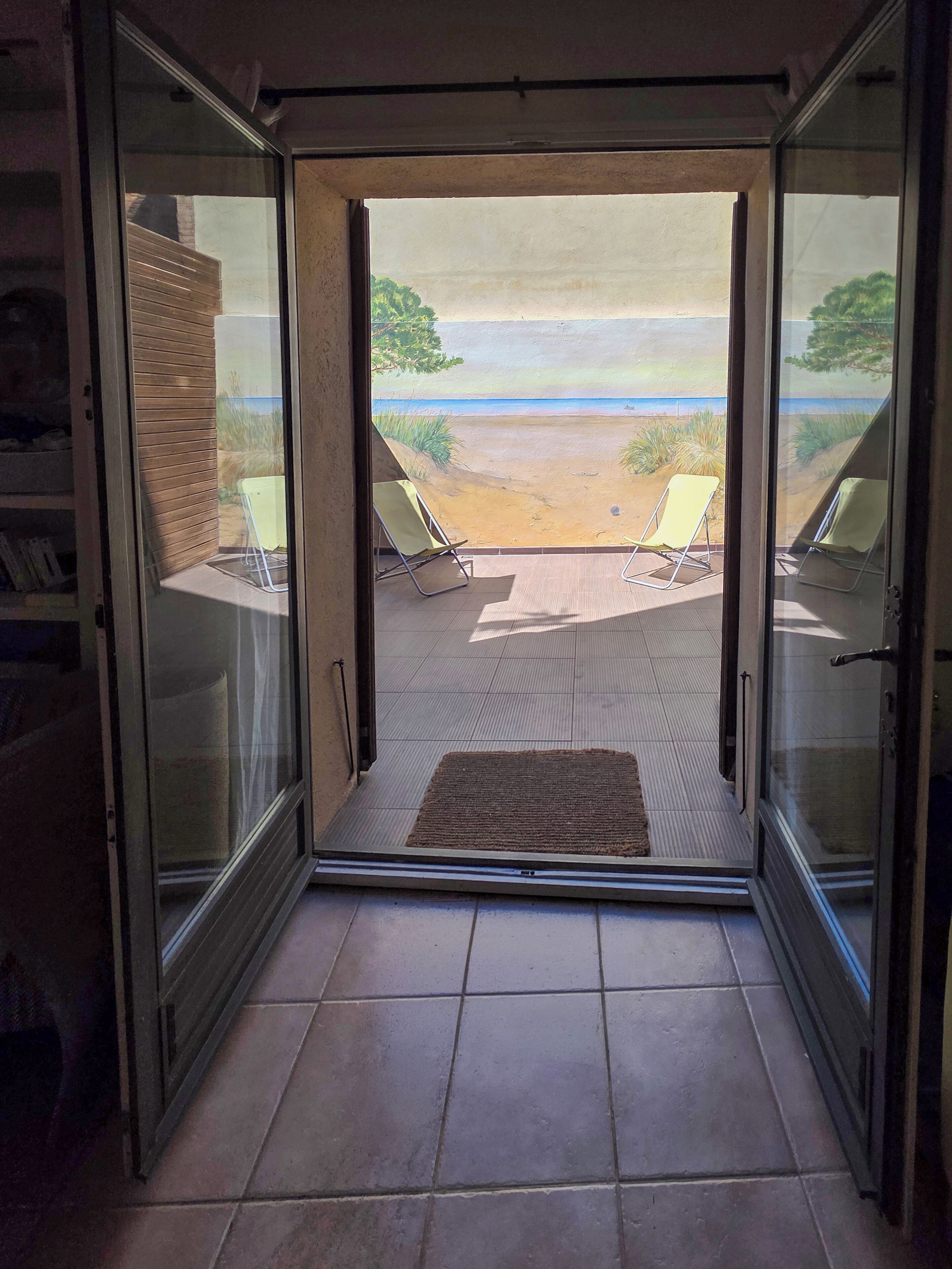 A glass door leading to a patio with two sun loungers and a view of a beach.