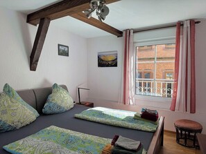 Een kamer met een bed, een raam met gordijnen, een klein krukje en een schilderij aan de muur.