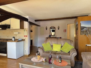 A compact living space with a kitchenette, a sofa with green cushions, and a dining table with glasses and bottles.