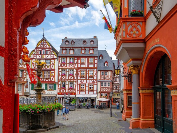 Uma praça de paralelepípedos com edifícios tradicionais em estilo enxaimel, uma fonte e áreas de estar ao ar livre.