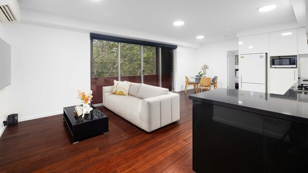 A modern living room with a sofa, a coffee table, and a kitchen area with a refrigerator and microwave. - Modern Comfort 2BR  Pool, Gym & Prime Valley Location (Fortitude Valley)
