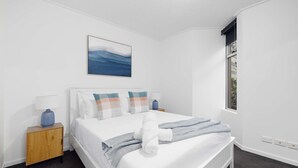 A neatly made bed with a blue and white checkered blanket, a blue vase on a wooden nightstand, and a painting of a blue ocean on the wall. - Modern Comfort 2BR  Pool, Gym & Prime Valley Location (Fortitude Valley)