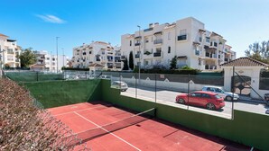 A tennis court with a green fence and a red surface, surrounded by residential buildings and parked cars. - Lovely 2-bed 10 Mins Walk From Puerto Banus (Marbella)