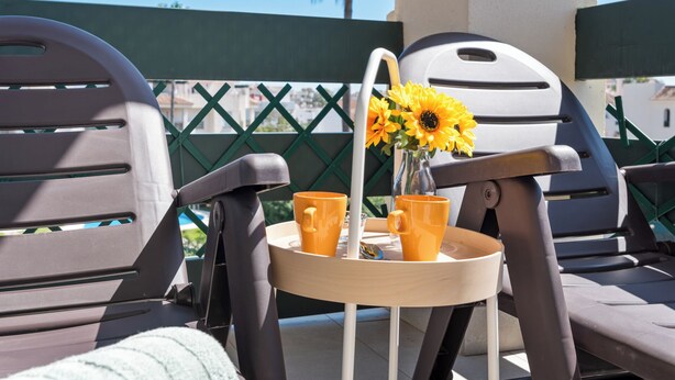 A sunlit outdoor seating area with two chairs, a small table with a vase of sunflowers and two orange cups, and a green fence in the background. - Lovely 2-bed 10 Mins Walk From Puerto Banus (Marbella)