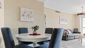 A modern living room with a round dining table, blue upholstered chairs, a grey sofa, and artwork on the walls. - Lovely 2-bed 10 Mins Walk From Puerto Banus (Marbella)