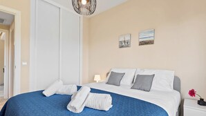 A neatly made bed with a blue quilt, white pillows, and two towels on the bed. There is a white nightstand with a lamp and a vase with a flower. The room has cream-colored walls and a door to the left. - Lovely 2-bed 10 Mins Walk From Puerto Banus (Marbella)