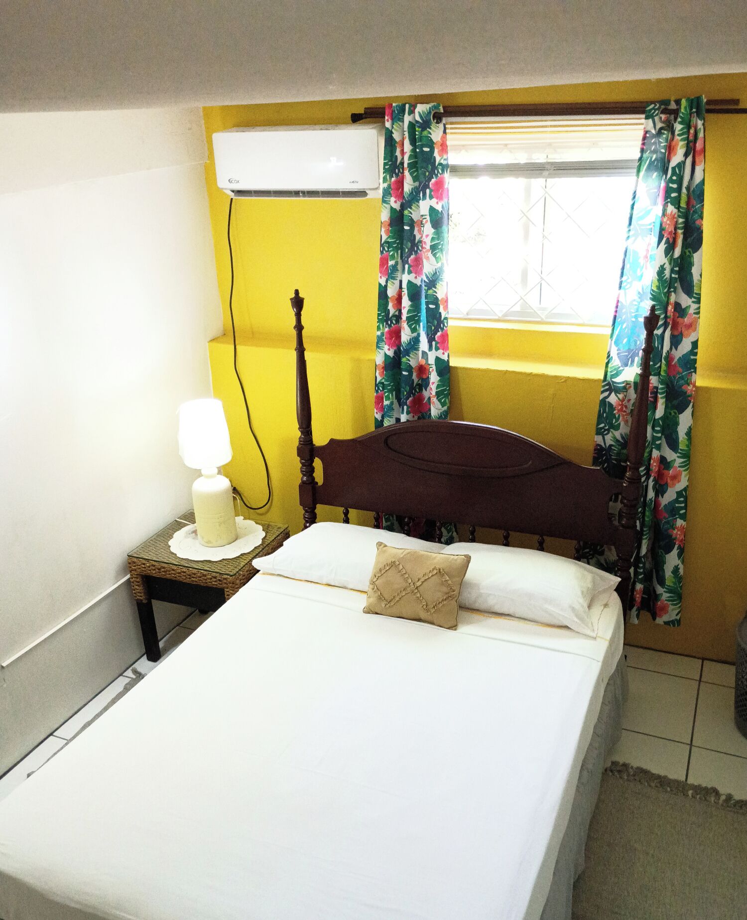 Una cama con cabecera de madera, una mesita de noche con lámpara y una ventana con cortinas florales.