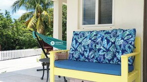 Un espace extérieur aménagé avec des sièges jaunes, agrémenté d’un coussin bleu orné d’un motif de feuilles tropicales, d’un hamac et, en arrière-plan, d’une clôture blanche en bois.