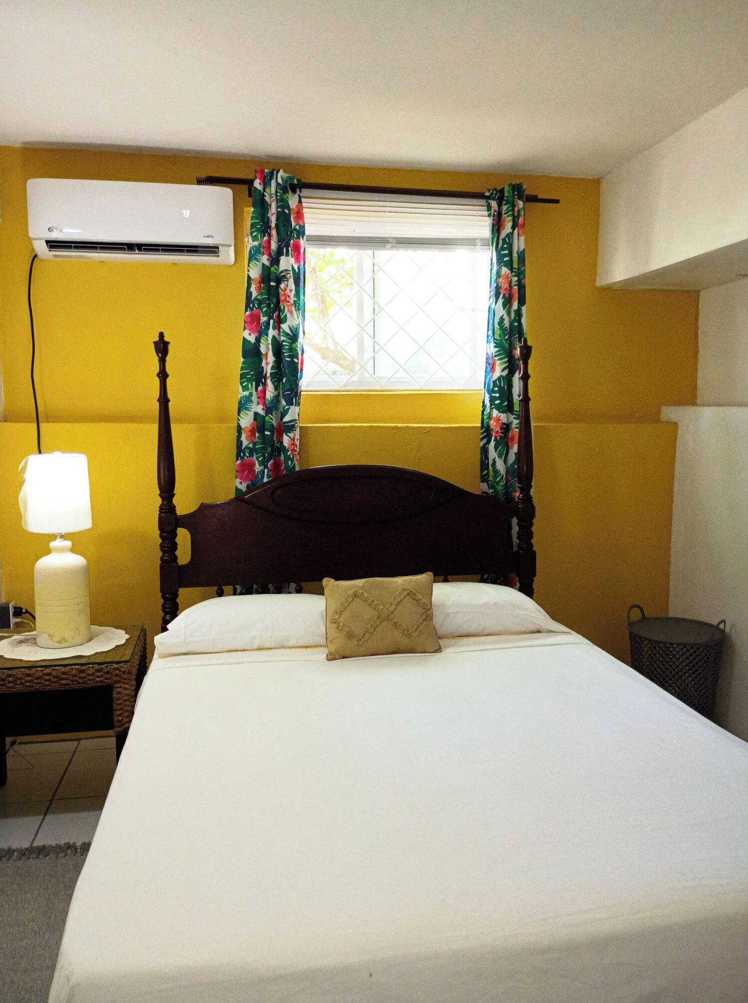 A bed with a wooden headboard, a nightstand with a lamp, and a window with floral curtains.