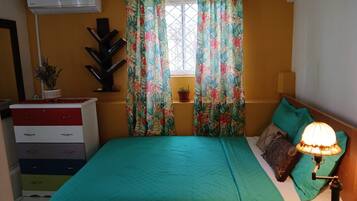 Un dormitorio con cama, lámpara de noche, ventana con cortinas, planta en maceta y aire acondicionado de pared.