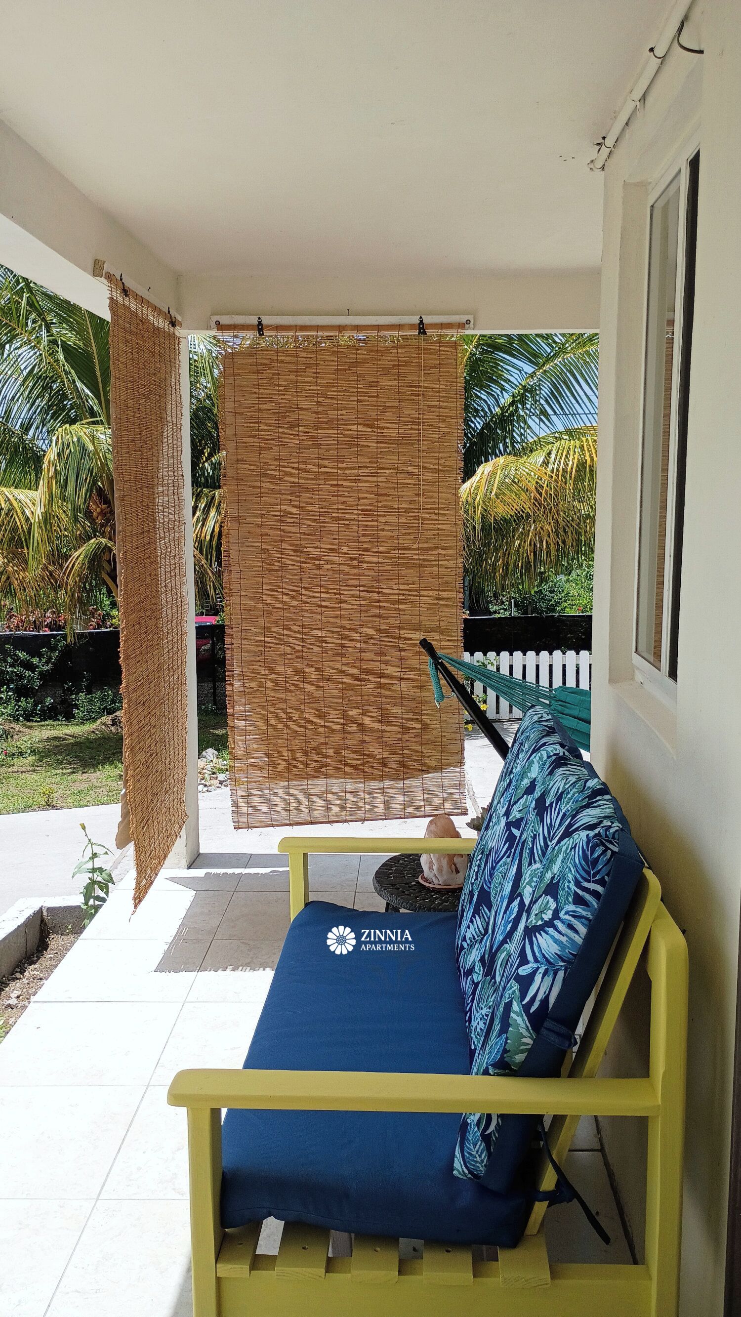 A covered outdoor seating area with a yellow bench and a blue cushion, a woven bamboo screen, and a white picket fence in the background.