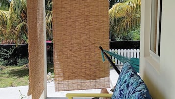 A covered outdoor seating area with a yellow bench and a blue cushion, a woven bamboo screen, and a white picket fence in the background.