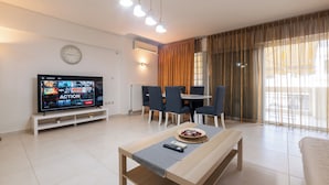 Ein modernes Wohnzimmer mit Flachbildfernseher, einem Essbereich mit Stühlen, einem Couchtisch und einem großen Fenster mit Vorhängen.