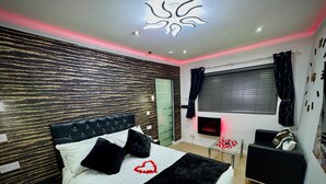 A modern bedroom with a tufted headboard, a bed with black and white bedding, a glass coffee table with red rose petals, a black speaker, and a wall-mounted TV. - Suite 1 Private Jacuzzi Spa Hot Tub & Sauna (Sheffield)