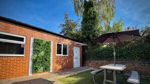 A brick building with a white door, a window with a shutter, and a small patio area with a table and bench under a large umbrella. - Suite 1 Private Jacuzzi Spa Hot Tub & Sauna (Sheffield)