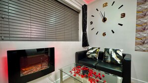 A modern living room with a fireplace, a glass coffee table with red decorations, a black sofa with patterned cushions, and a wall clock. - Suite 1 Private Jacuzzi Spa Hot Tub & Sauna (Sheffield)