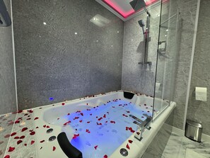 Ein modernes Badezimmer mit einer Glaskonstruktion als Badewanne, gefüllt mit Rosenblättern und Kerzen, Marmorwänden und einer Duschkabine.