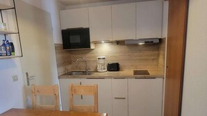 Eine kleine Küche mit weißen Schränken, einer Mikrowelle, einer Kaffeemaschine und einem Toaster. Ein Holztisch und zwei Stühle stehen bereit.
