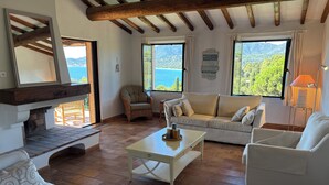 A cozy living room with a fireplace, comfortable seating, and large windows offering a view of the sea.