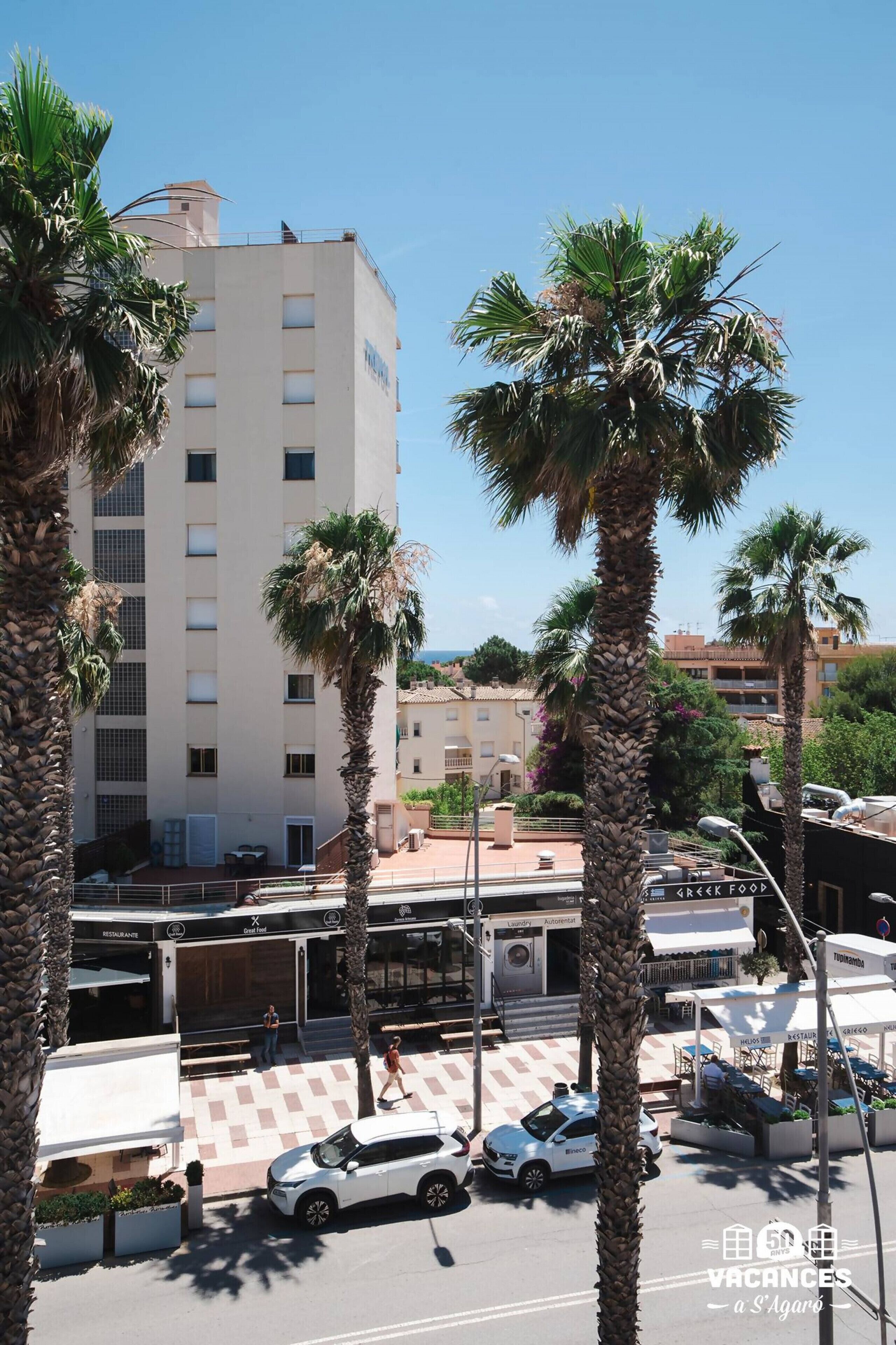 A multi-story building with a flat facade, palm trees in front, and a street with cars and pedestrians.