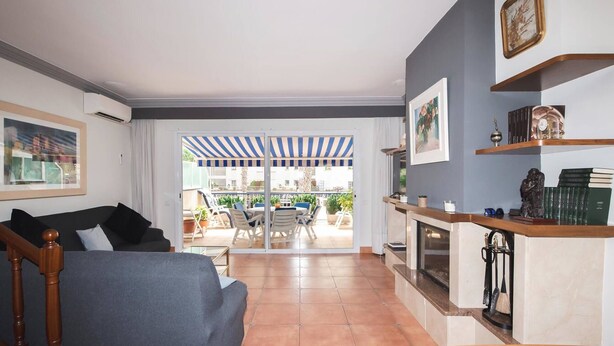 Un salon avec un canapé, une table basse et une cheminée. Il y a un balcon avec un auvent rayé et une vue sur la ville.