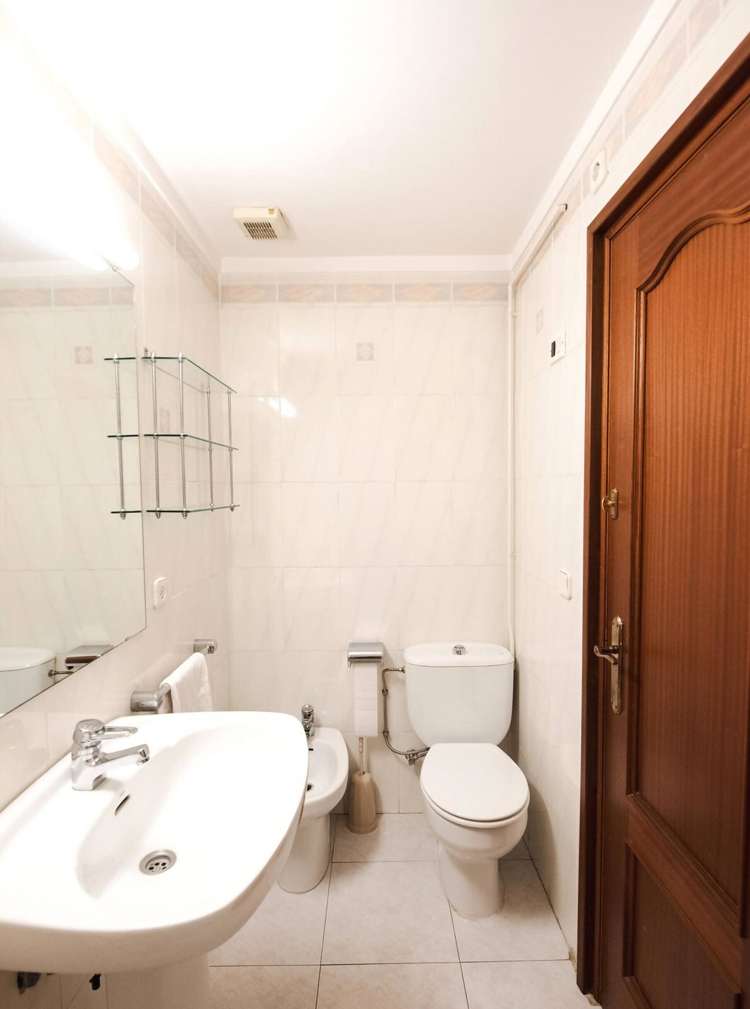 Een badkamer met een wastafel, toilet en bidet. Er is een houten deur en een spiegel aan de muur.