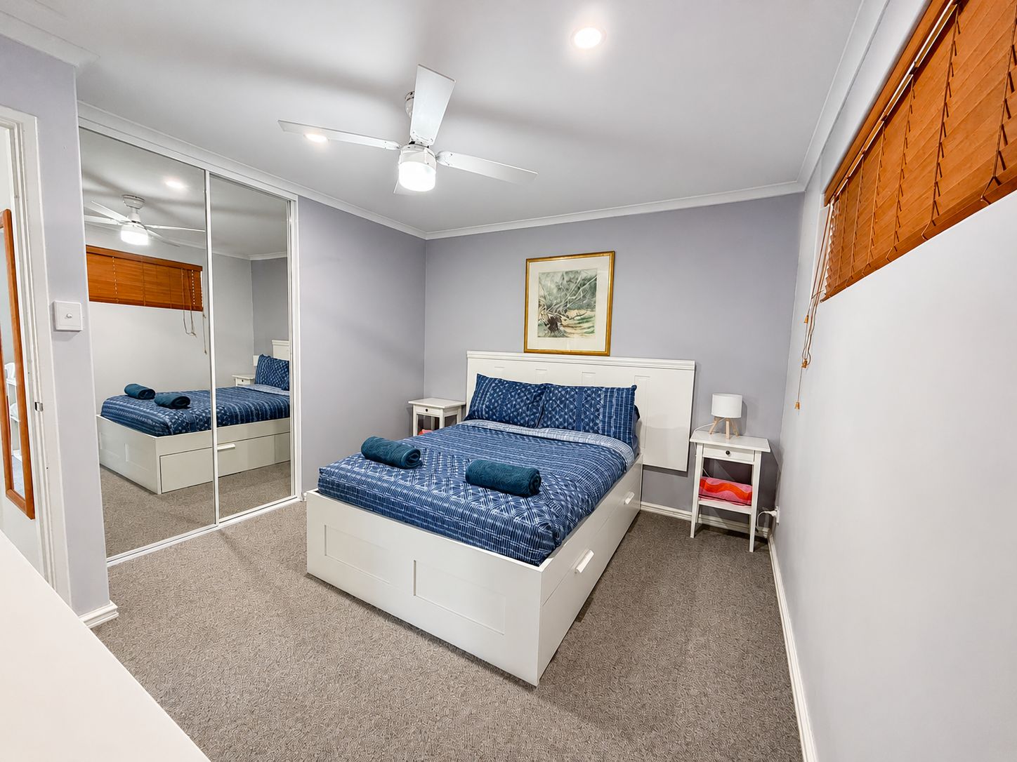 A bedroom with a bed, a ceiling fan, a bedside table, a nightstand, a mirror, and a window with blinds.