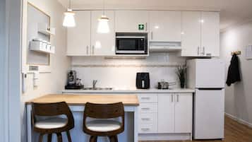 A modern kitchen with white cabinets, a microwave, a coffee machine, and a refrigerator. There are two wooden bar stools and pendant lights above the counter.