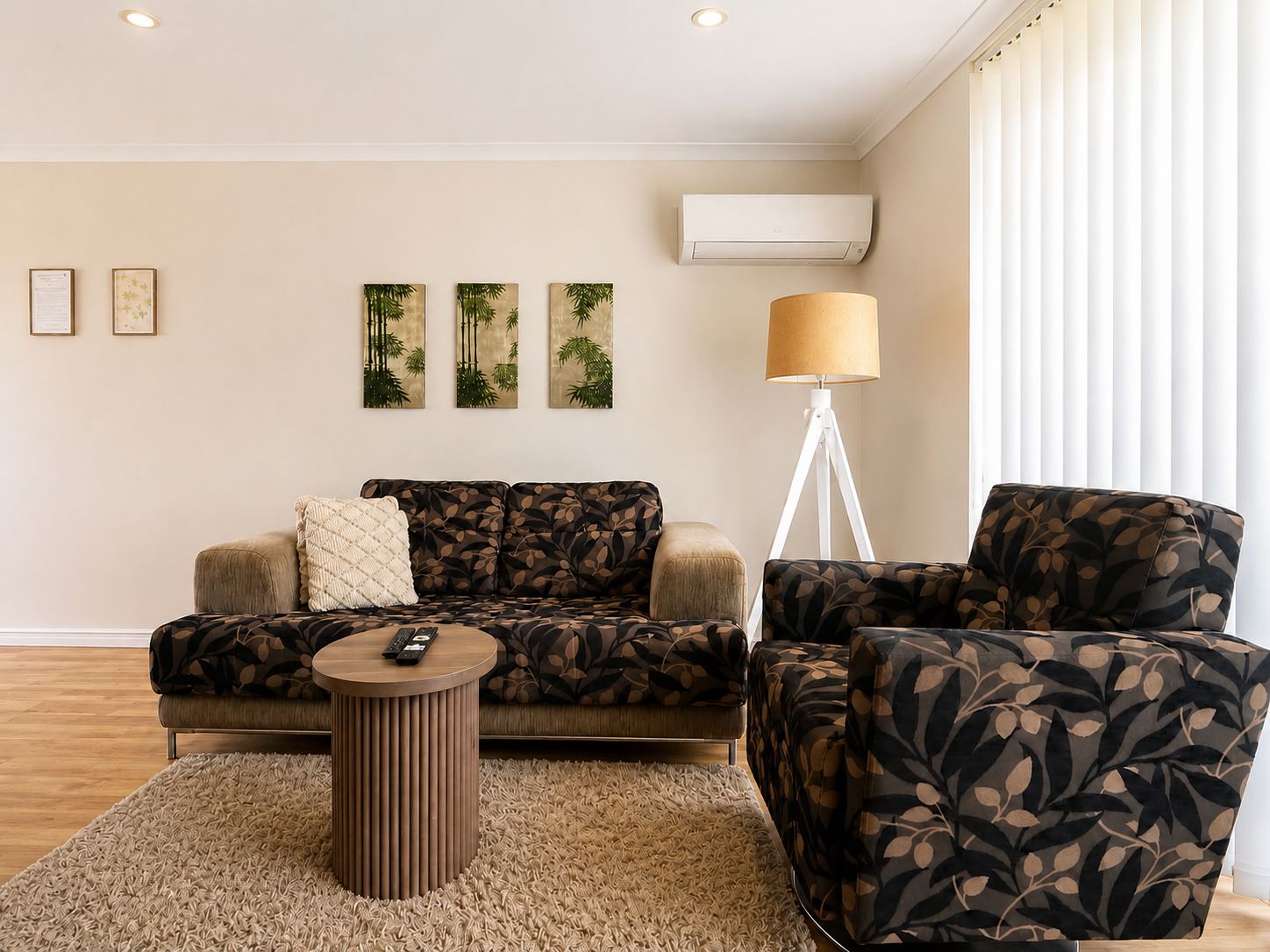 A living room with a sofa, armchair, and a small round table. There are three wall-mounted paintings, a floor lamp, and an air conditioning unit.