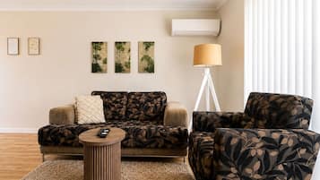 A living room with a sofa, armchair, and a small round table. There are three wall-mounted paintings, a floor lamp, and an air conditioning unit.
