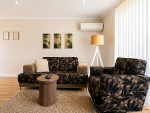 A living room with a sofa, armchair, and a small round table. There are three wall-mounted paintings, a floor lamp, and an air conditioning unit. - Josh's Sea Breeze | Beach Walk, Pool & Marina (Mandurah)