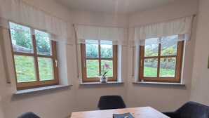 Una habitación con una mesa de madera y cuatro sillas negras, un florero con flores en el alféizar de la ventana y una lámpara en el techo.
