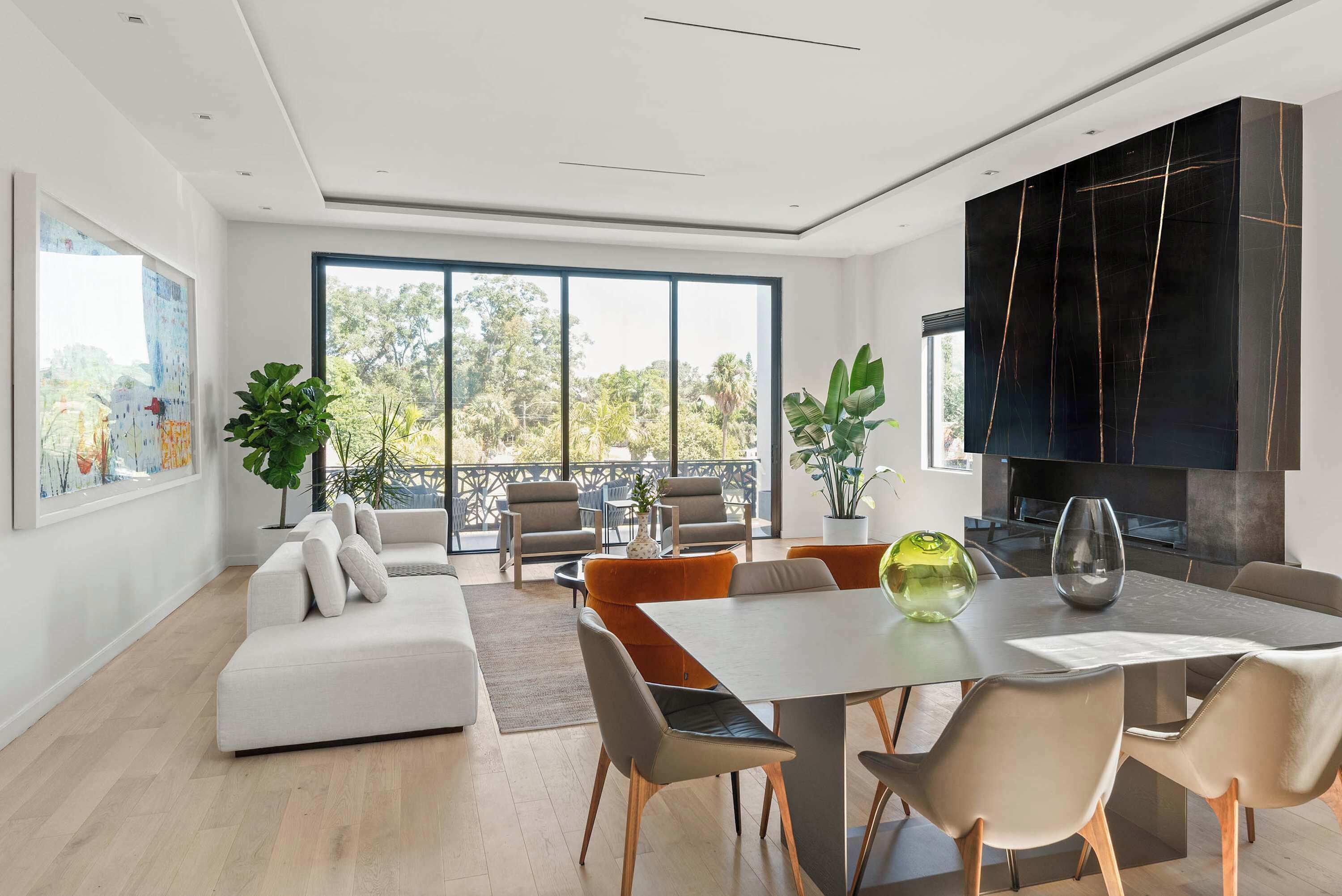 A modern living room with a white sofa, orange chairs, a dining table, and large windows offering a view of greenery.