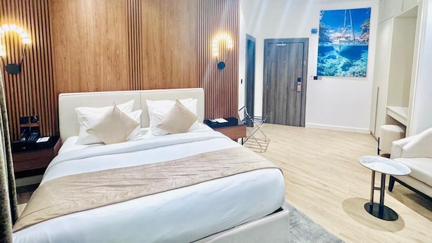 A modern hotel room with a large bed, wooden paneling on the walls, a painting of a boat on the wall, and a small round table. - ATLANTIS HOTELS (Lagos)