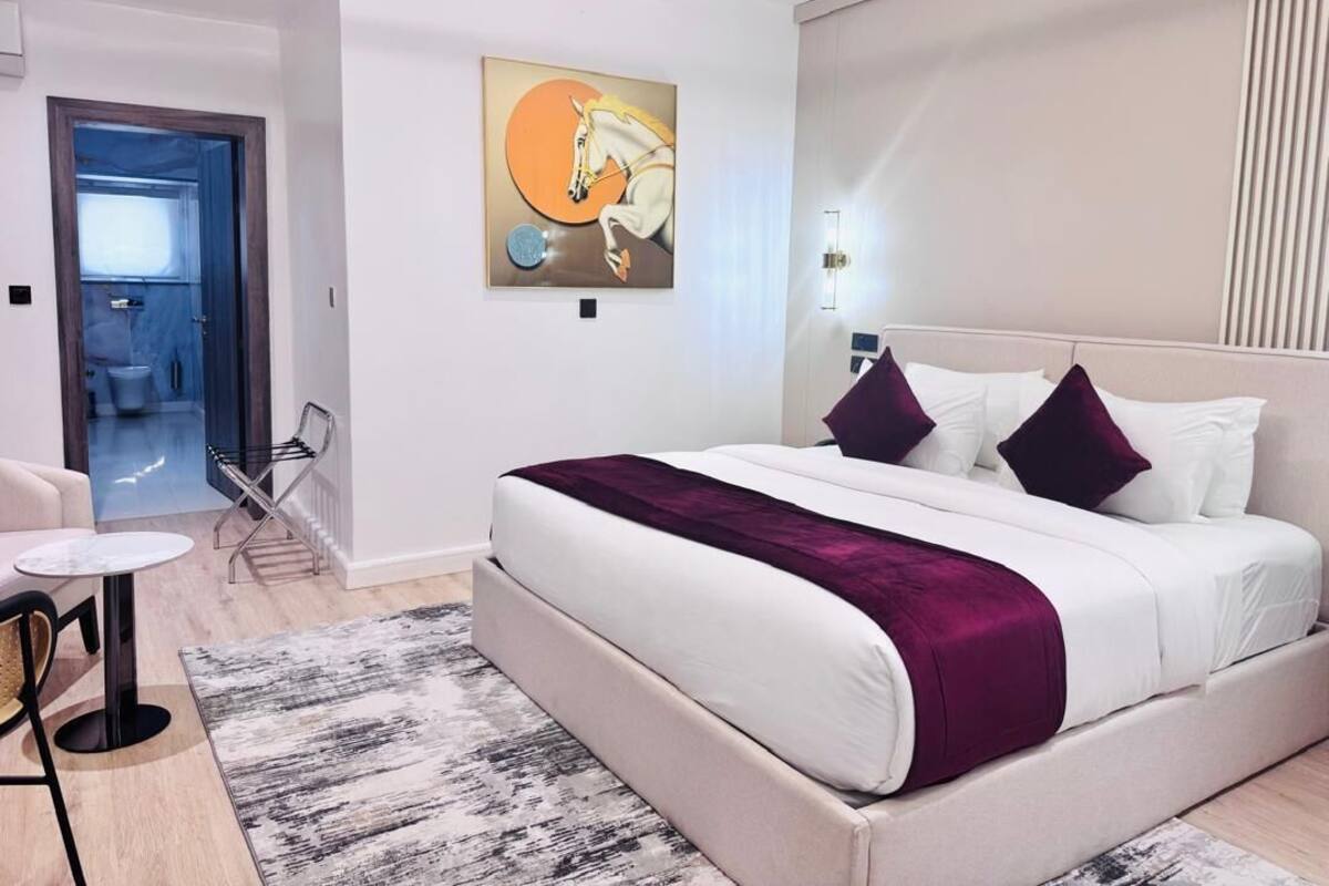 A modern bedroom with a large bed, a small table, and a chair. There is a painting on the wall and a door leading to another room.