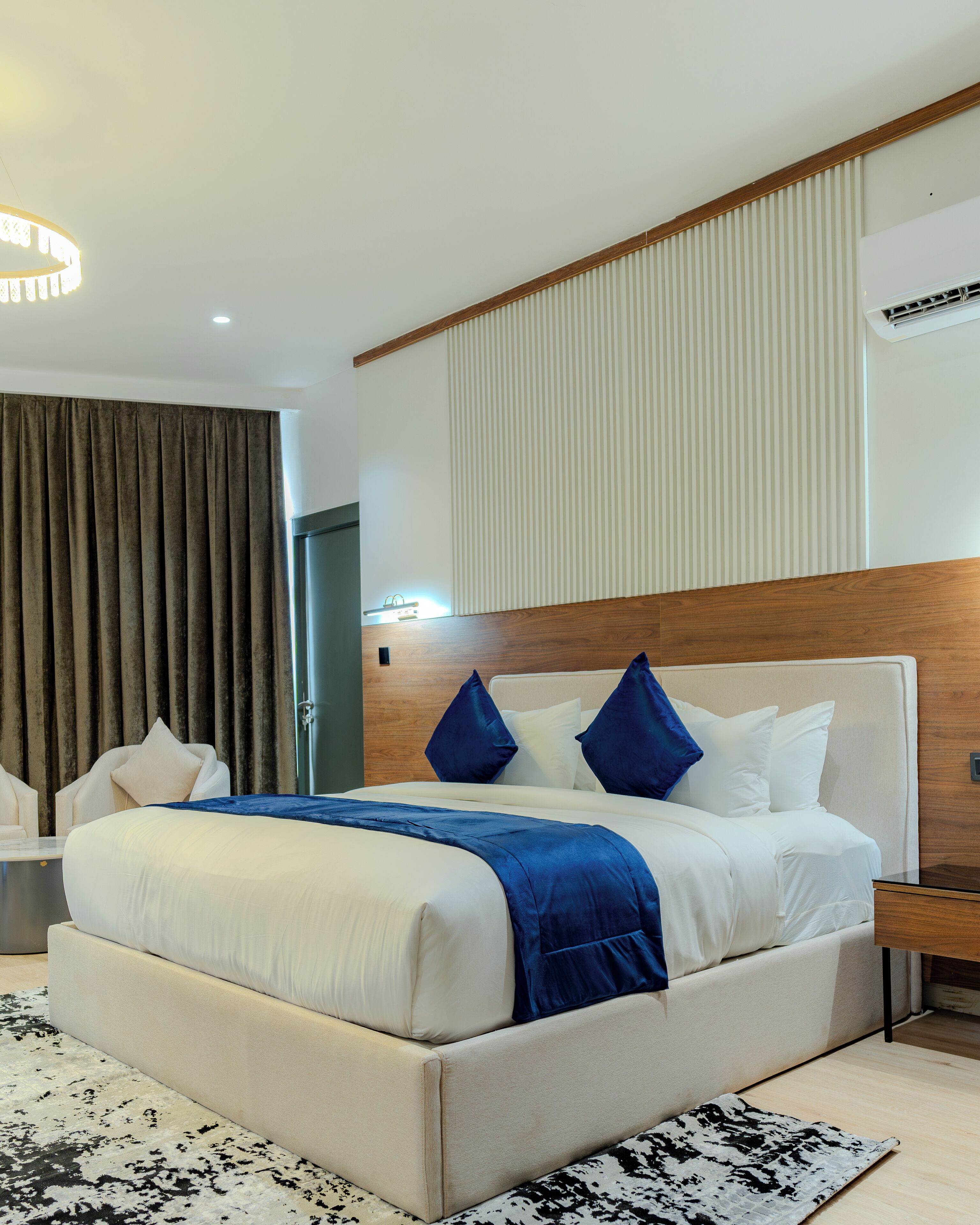 Une chambre d’hôtel moderne avec un grand lit, une tête de lit en bois, une table de chevet, une commode, un bureau et une grande fenêtre avec des rideaux.