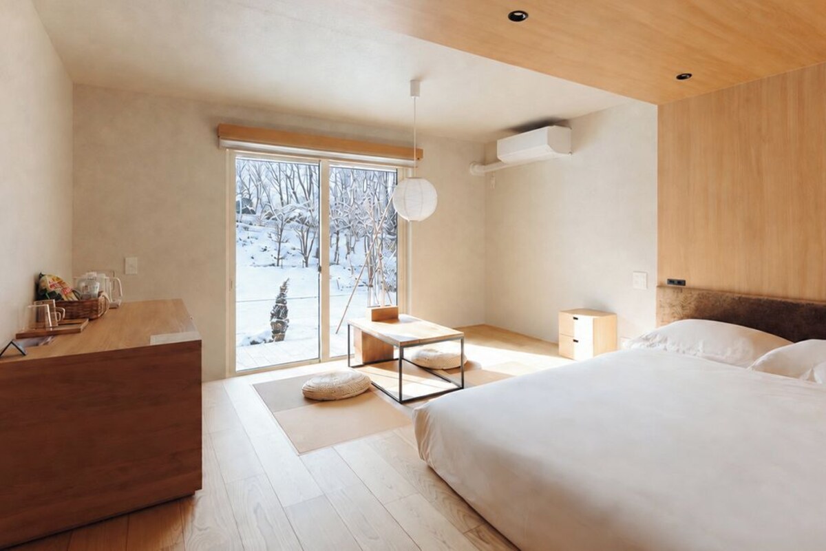 A modern bedroom with a large bed, a wooden dresser, a desk, and a view of a snowy landscape through the window.