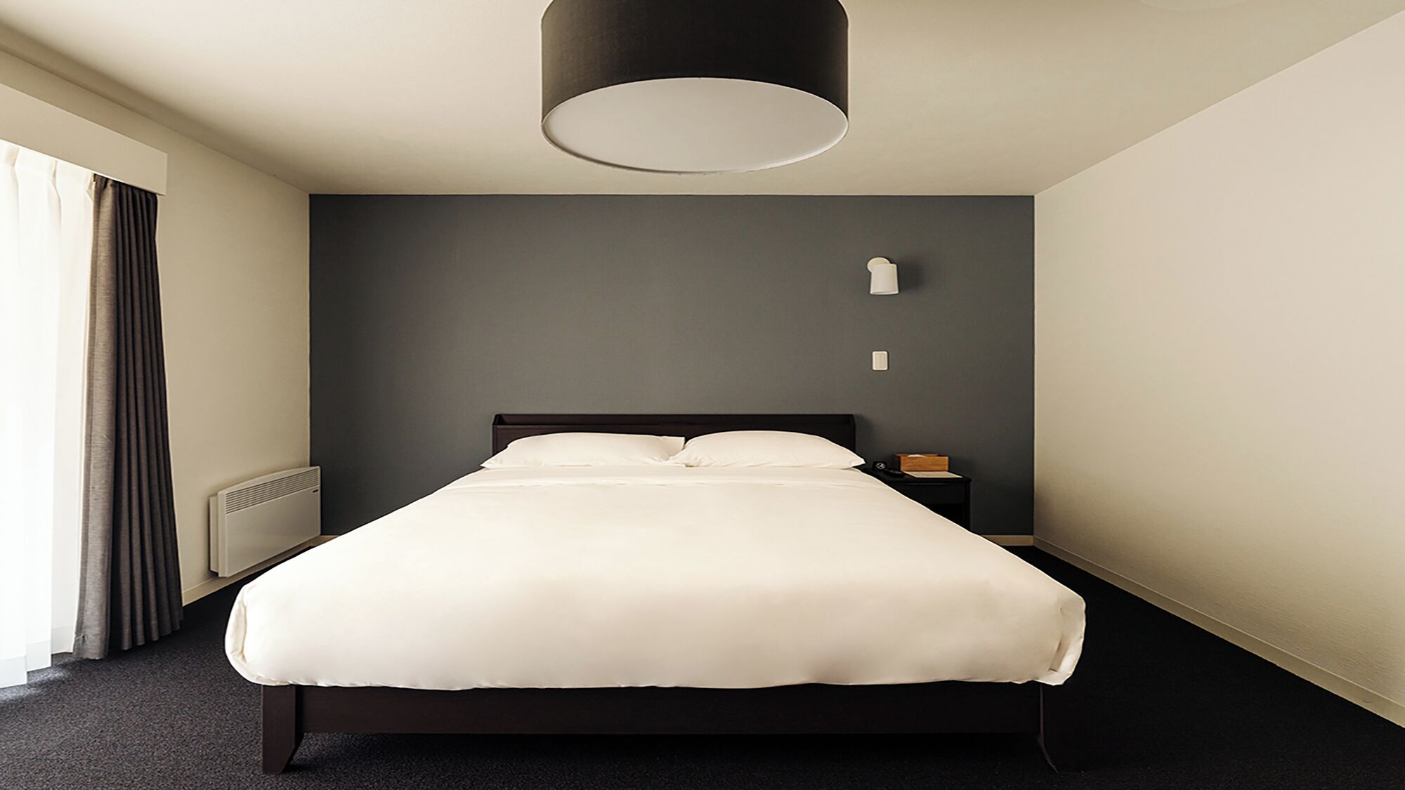 A modern hotel room with a large bed, a bedside table, a lamp, and a window with curtains.