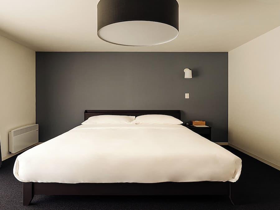 Una habitación de hotel moderna con una cama grande, una mesita de noche, una lámpara y una ventana con cortinas.