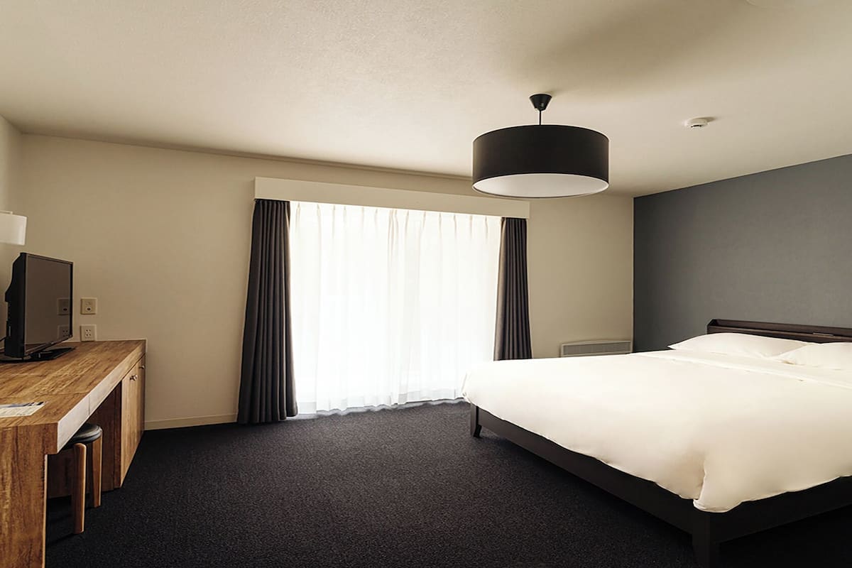 Habitación de hotel con cama, escritorio con computadora, lámpara y ventana con cortinas.